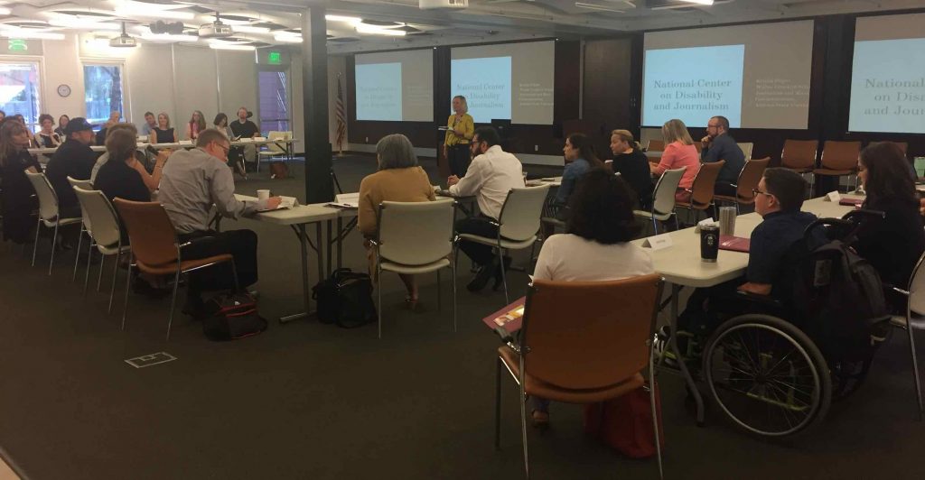 NCDJ Director Kristin Gilger introduces participants and event coordinators at the start of the April 25, 2018 "Improving Disability Communication" workshop at Ability 360. About 30 workshop attendees sit around a horseshoe-shaped table facing three large projection screens.